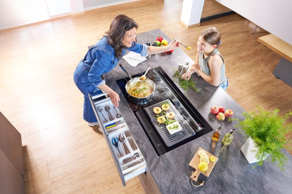 Eine Frau kocht gemeinsam mit einem Mädchen an einer Kücheninsel mit integriertem ORANIER-Kochfeldabzug. Dampf wird leise und direkt aus der Mitte des Kochfelds abgesaugt – offene Wohnküche mit ruhiger Atmosphäre.